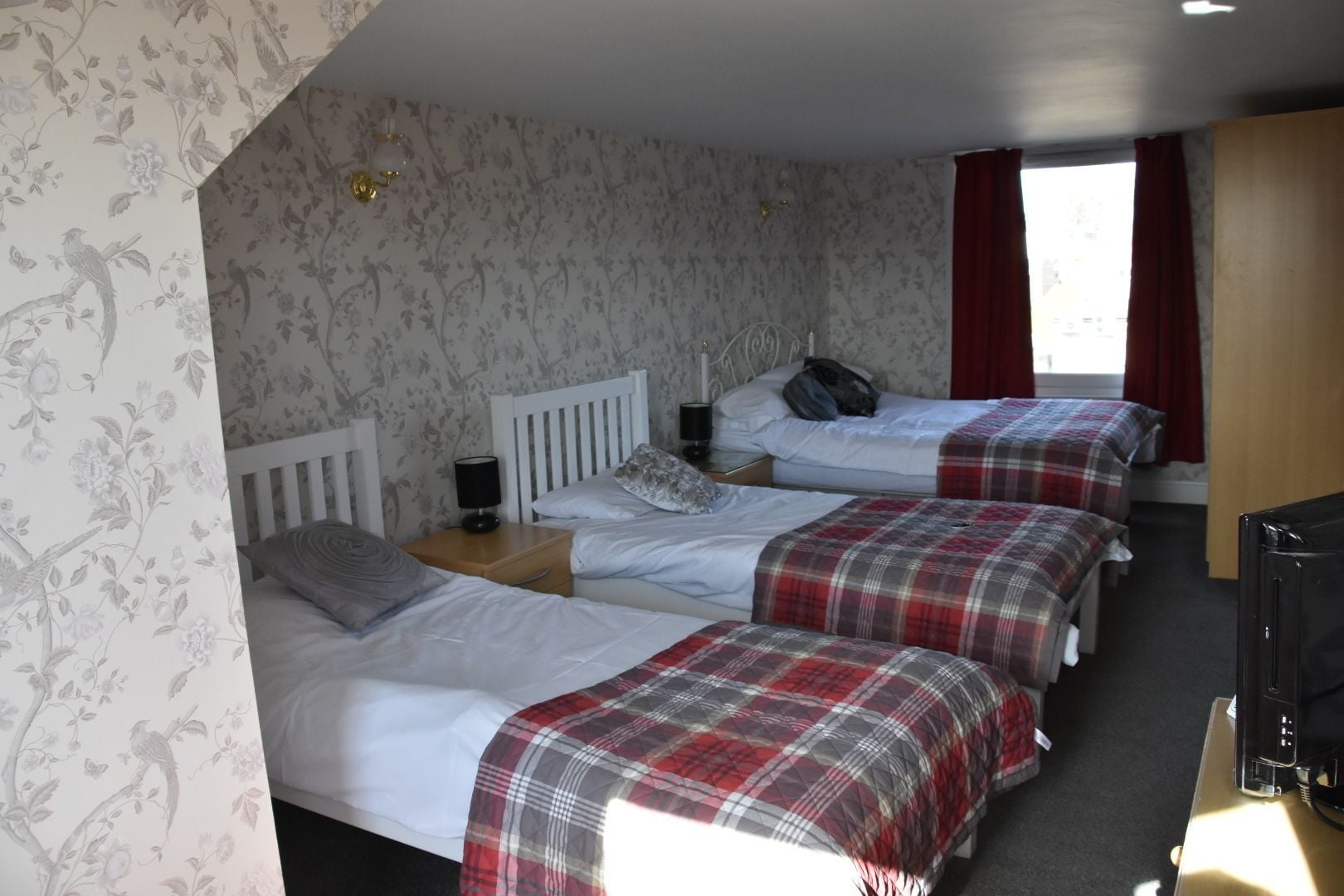 Une chambre avec trois lits, chacun recouvert d’une couverture à carreaux rouges et gris, avec du linge de lit blanc et une tête de lit blanche. On y trouve une petite table avec une lampe et un téléviseur posé sur un meuble. Les murs sont recouverts d’un papier peint à motifs floraux et il y a une fenêtre avec des rideaux rouges.