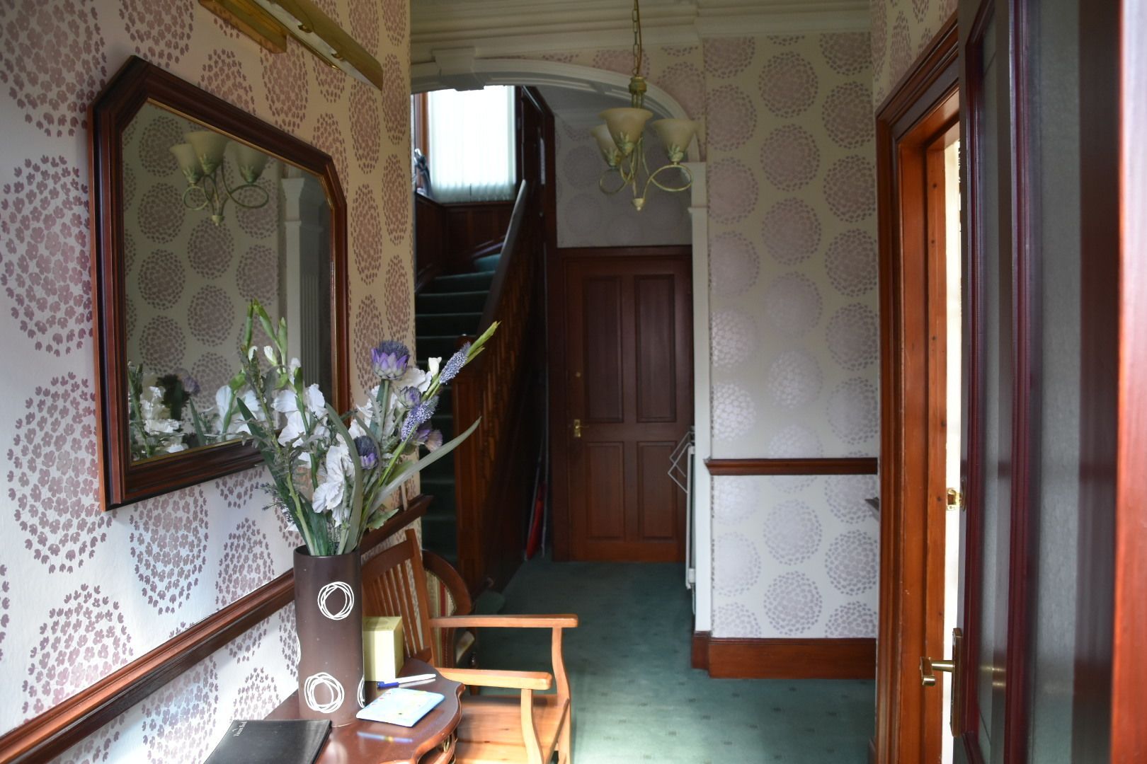 A hallway with patterned wallpaper, a wooden door, a staircase, and a mirror with flowers.