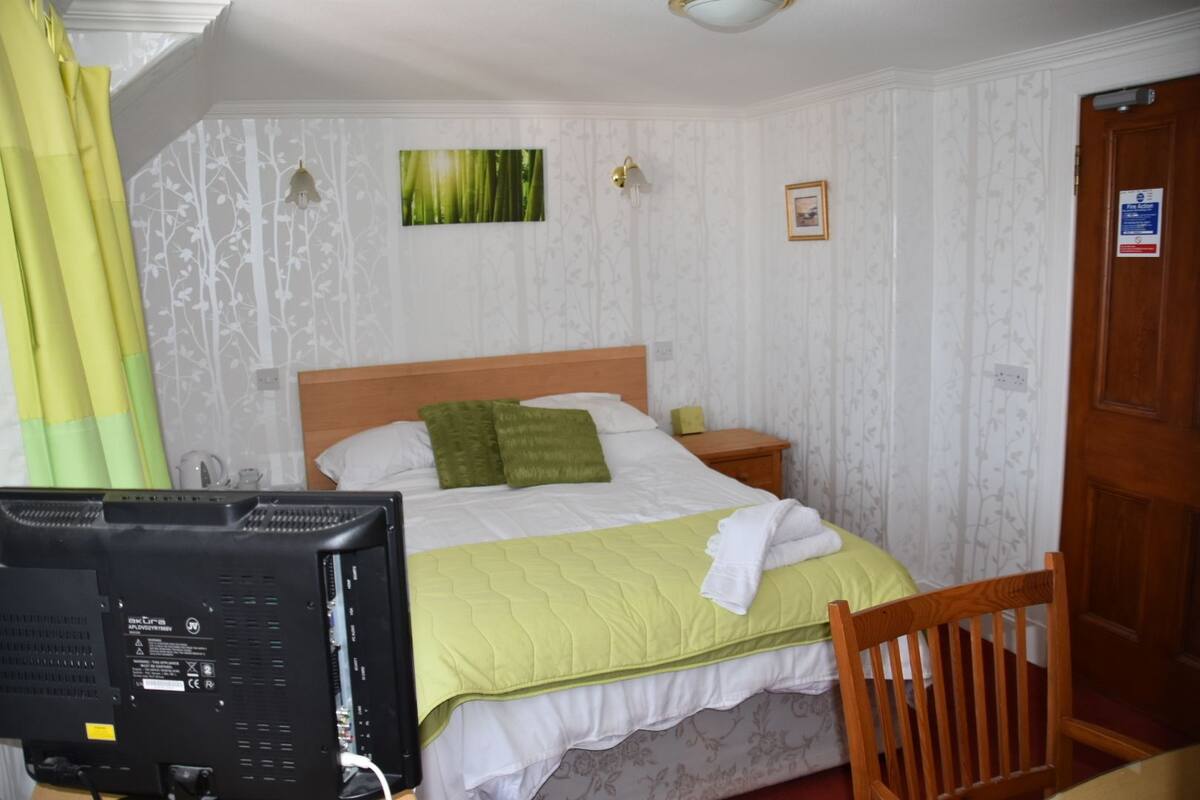 A hotel room with a bed, a desk with a computer, a chair, a door, and a window with curtains.