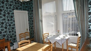 A dining area with a wooden table and chairs, a white tablecloth, and a window with vertical blinds. - The Gables (Bathgate)