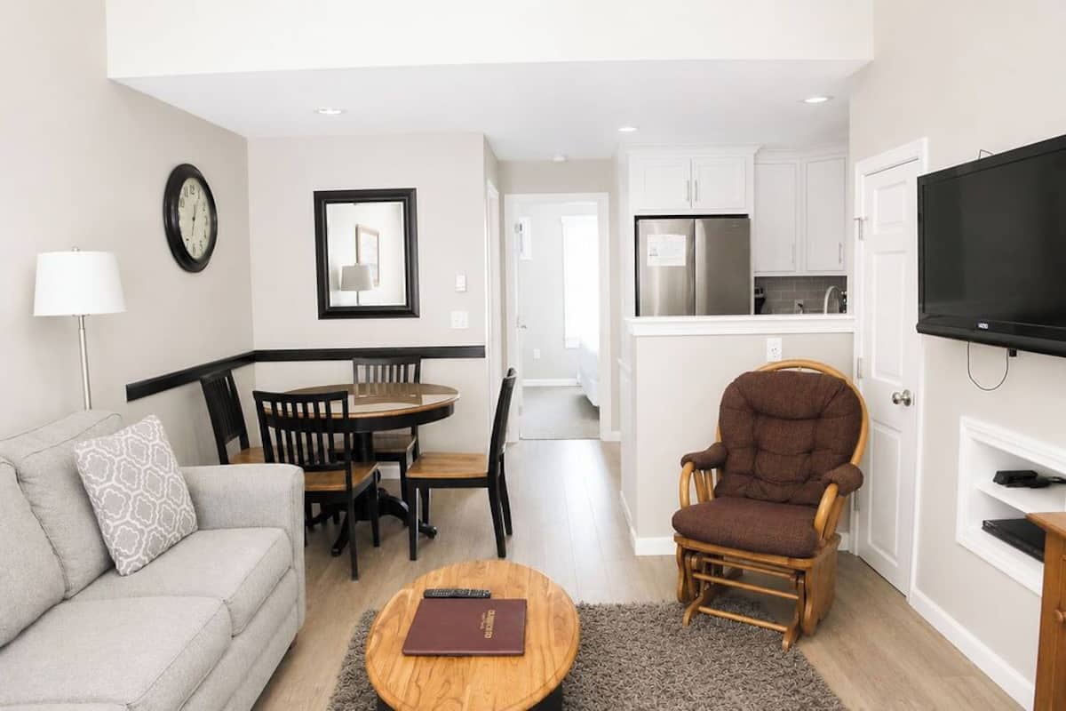 A living room with a sofa, a round coffee table, a dining table with chairs, a television, and a kitchen area with cabinets and a refrigerator.