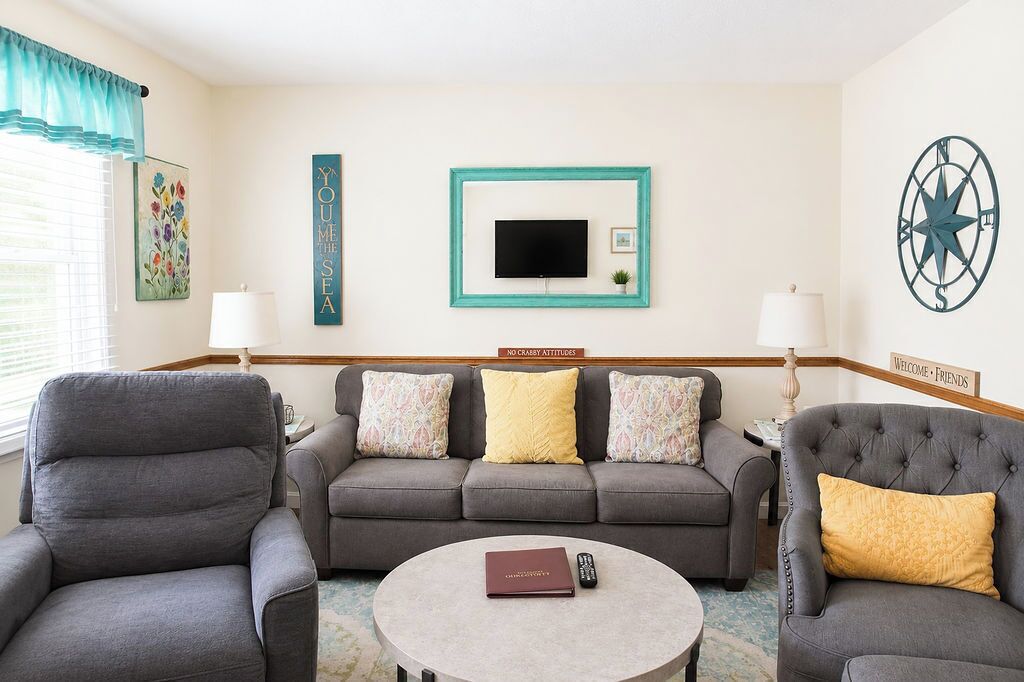 A room with a grey sofa, two armchairs, a round coffee table, a TV, and wall decorations including a compass and signs.