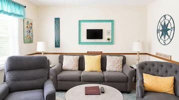 A room with a grey sofa, two armchairs, a round coffee table, a TV, and wall decorations including a compass and signs.