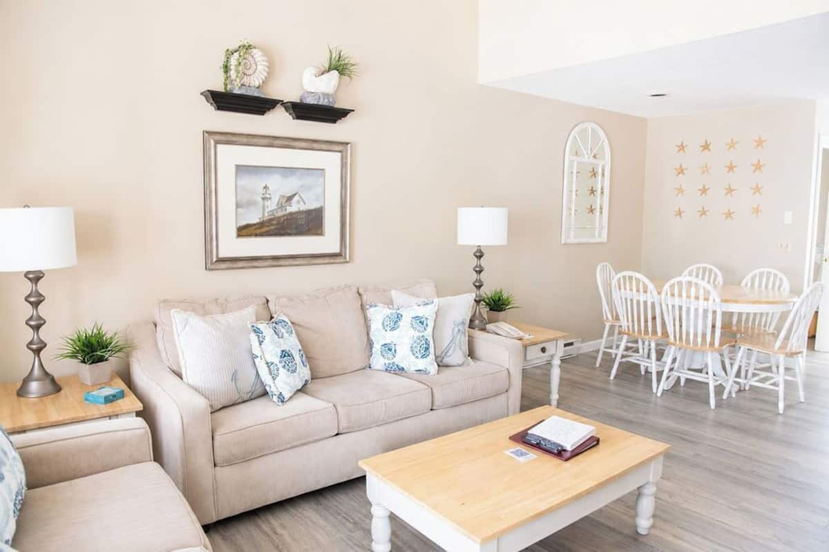 A living room with a beige sofa, a wooden coffee table, a dining table with white chairs, a framed picture on the wall, and a small plant on the side table.