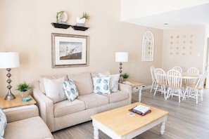 A living room with a beige sofa, a wooden coffee table, a dining table with white chairs, a framed picture on the wall, and a small plant on the side table.