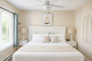 A bedroom with a bed, two bedside tables with lamps, a ceiling fan, a sailboat artwork on the wall, and a window with curtains. - Elmwood Resort Hotel (Wells)