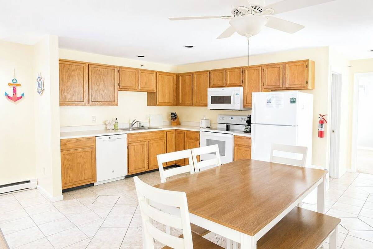 A kitchen with wooden cabinets, a white countertop, a dining table with chairs, a ceiling fan, and a white refrigerator.