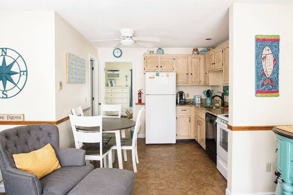 A compact kitchen with wooden cabinets, a white refrigerator, and a dining area with a gray sofa and white chairs.