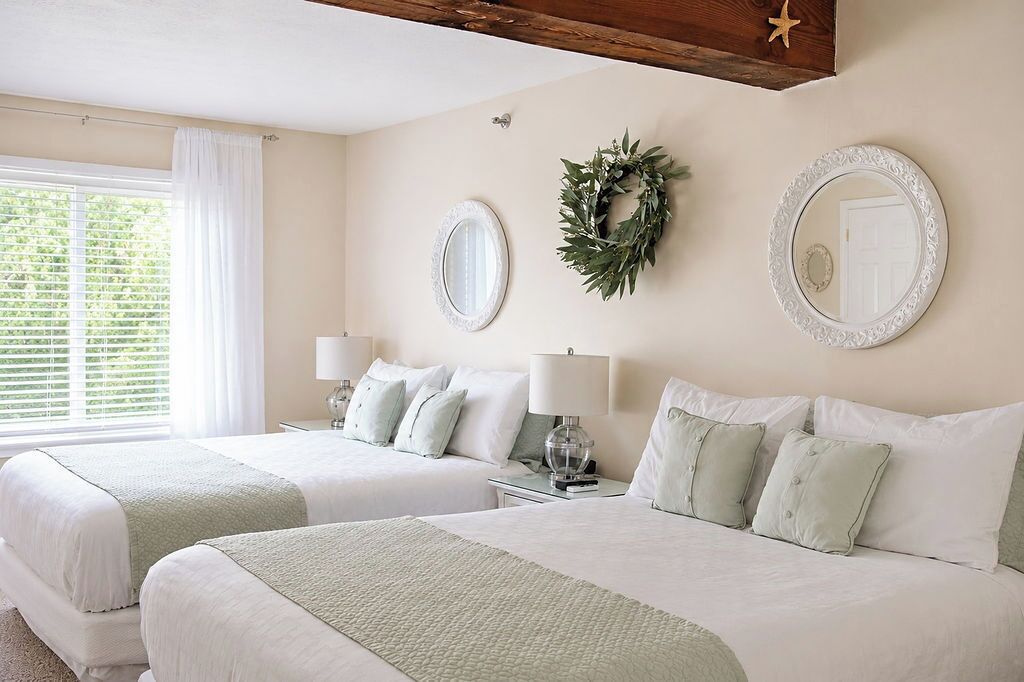 A hotel room with two beds, a window with white curtains, a wall-mounted mirror, and a wreath.