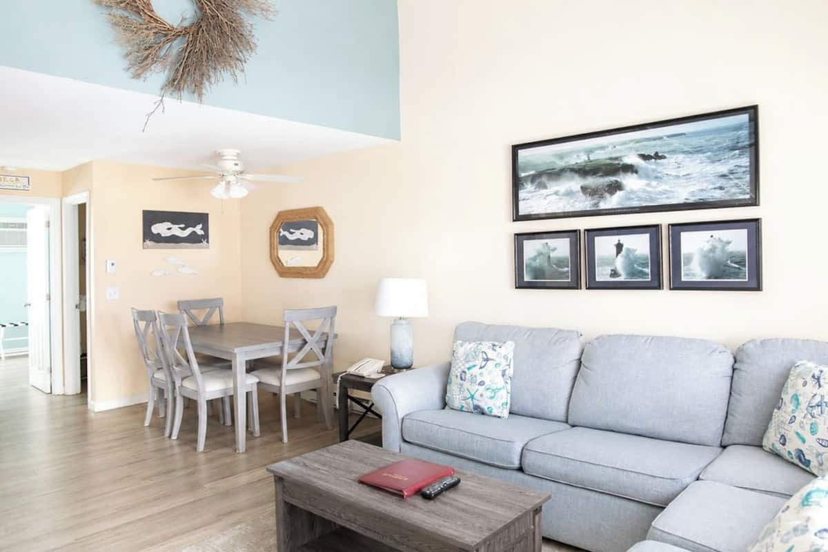 A living room with a grey sofa, a dining table with chairs, a coffee table, and framed pictures on the wall.