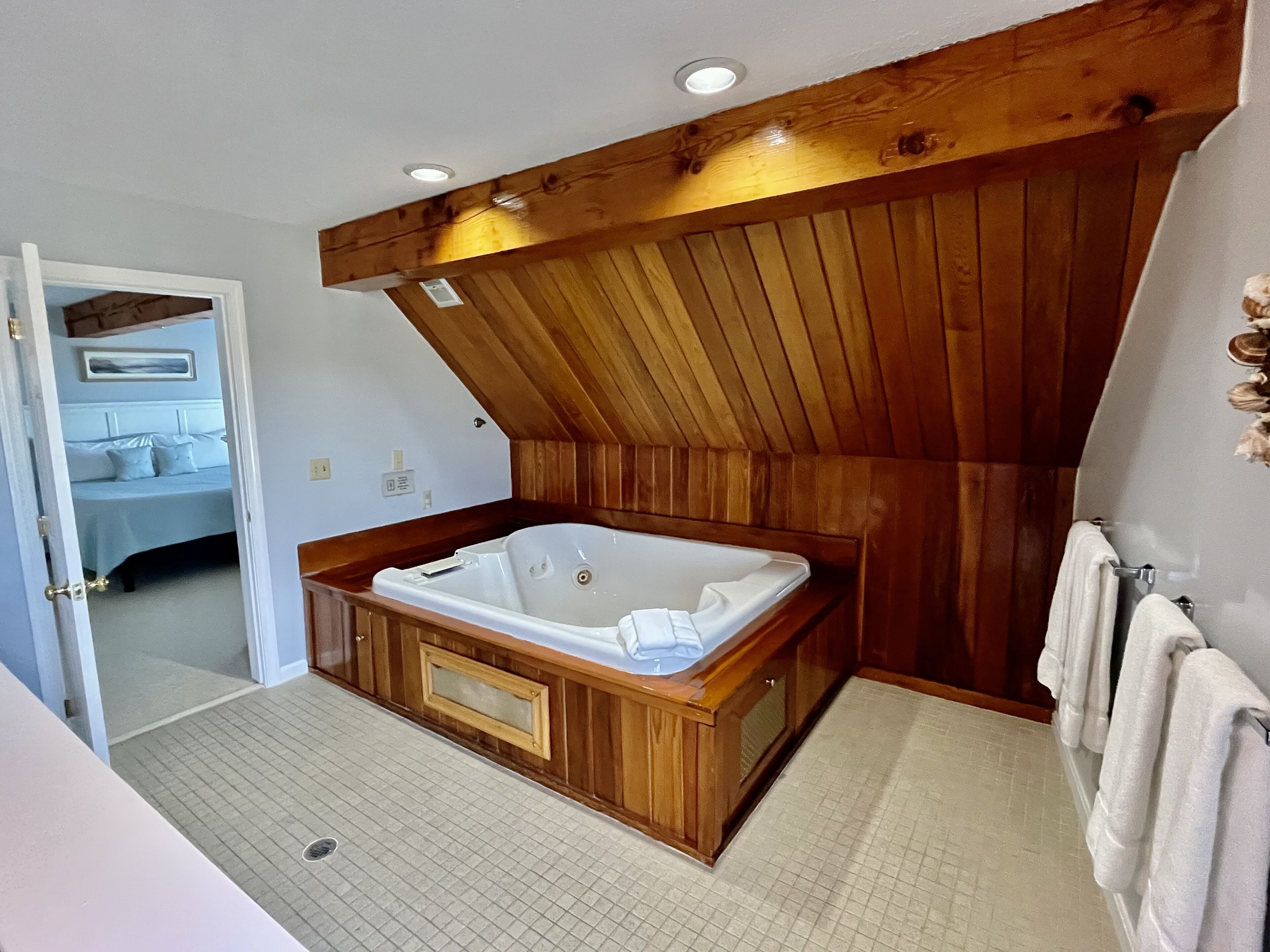 A bathroom with a wooden bathtub, a shower area, and a visible bedroom with a bed and a painting on the wall.