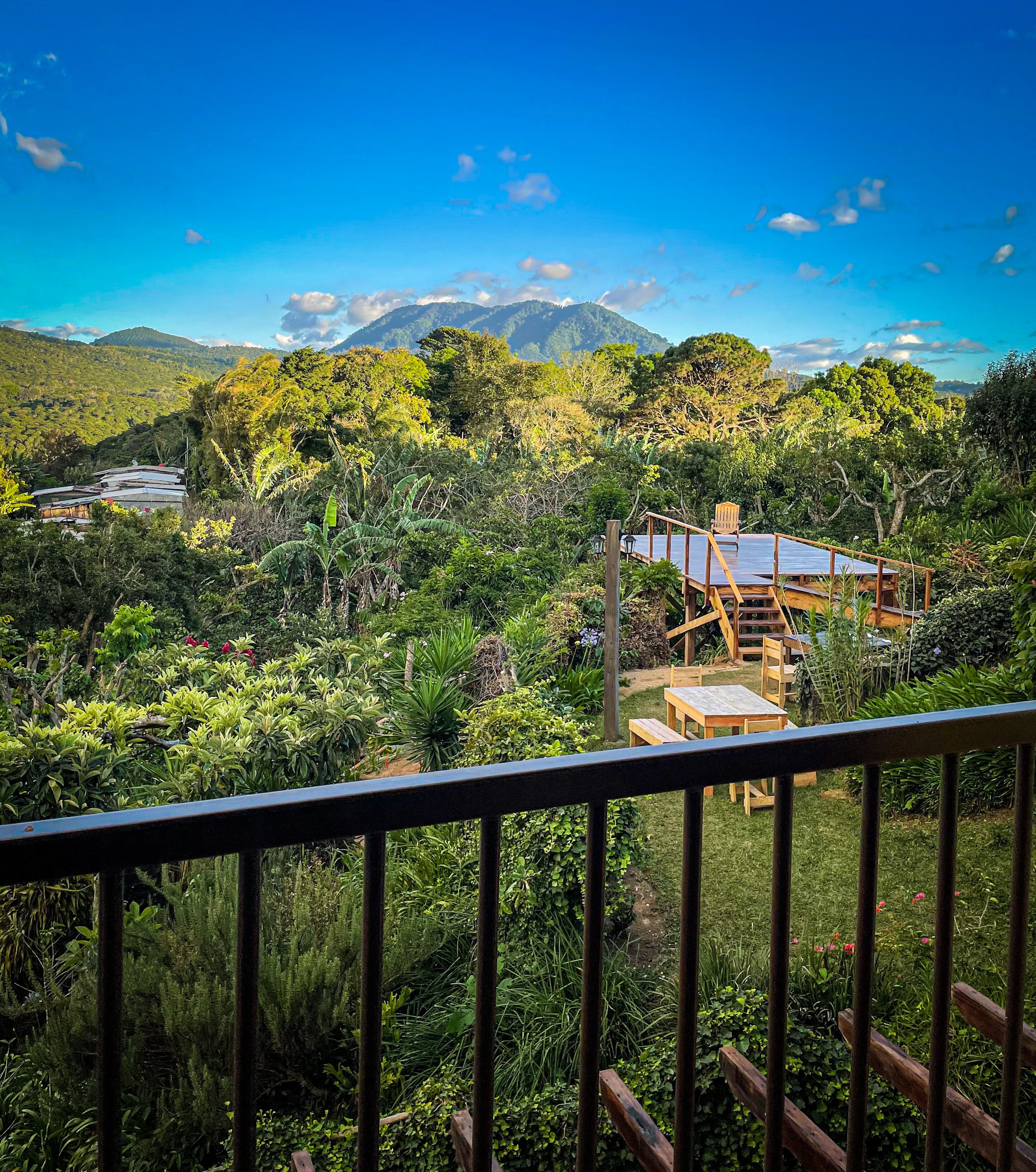 A view from a balcony overlooking a lush garden with a wooden deck, a mountain in the background, and a clear blue sky.