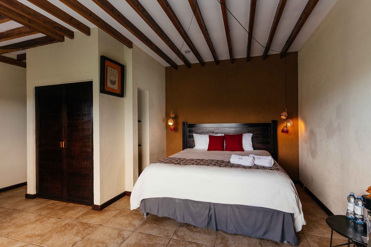 Ein Hotelzimmer mit einem großen Bett, Holzbalkendecke, einer dunklen Tür und einem gerahmten Bild an der Wand.