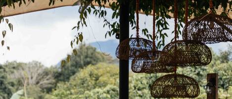 An outdoor dining area with wooden tables and metal chairs, surrounded by lush greenery and a view of hills.