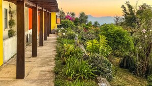 Une allée couverte, avec des poutres en bois, mène à un bâtiment aux murs jaunes et oranges, entouré d’une végétation luxuriante et offrant une vue sur des montagnes lointaines.