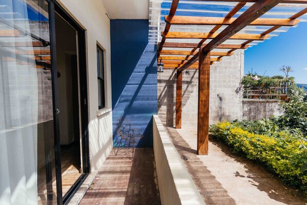 Ein schmaler Außenbereich mit einer Holz-Pergola, einem Metallstuhl und Blick auf einen Garten mit gelben Blumen.