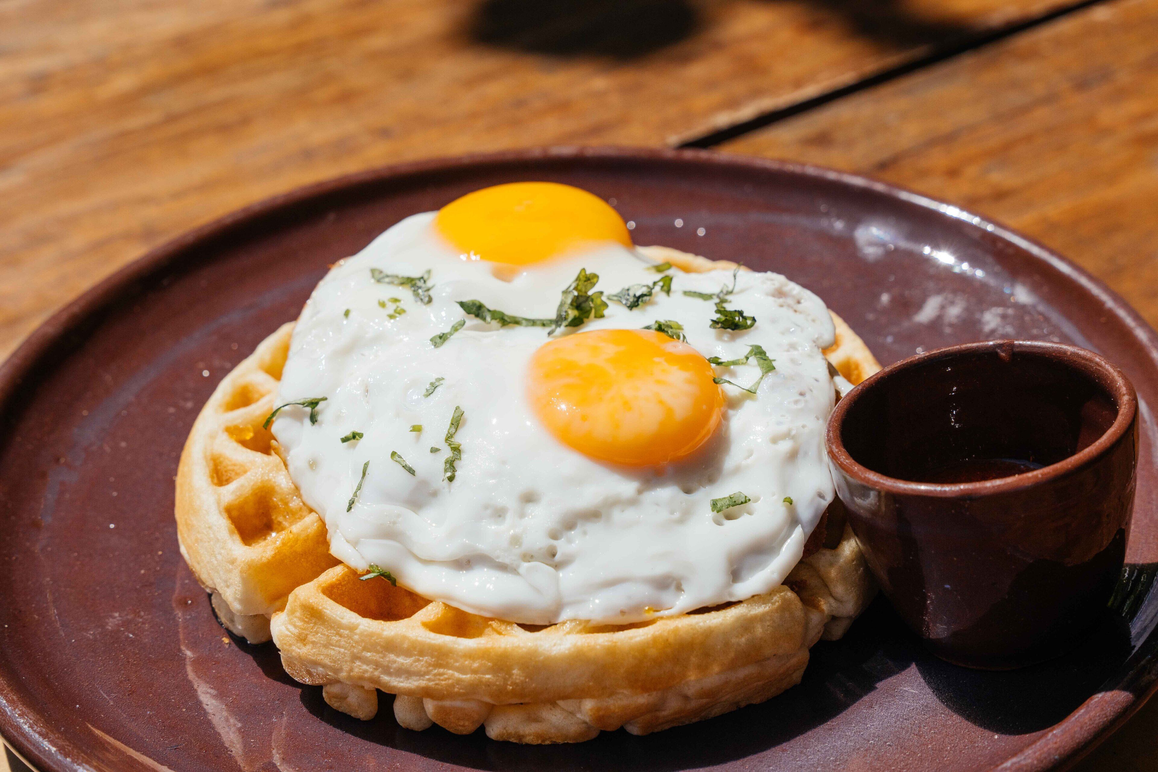 A waffle topped with two sunny-side-up eggs and a small cup of syrup.