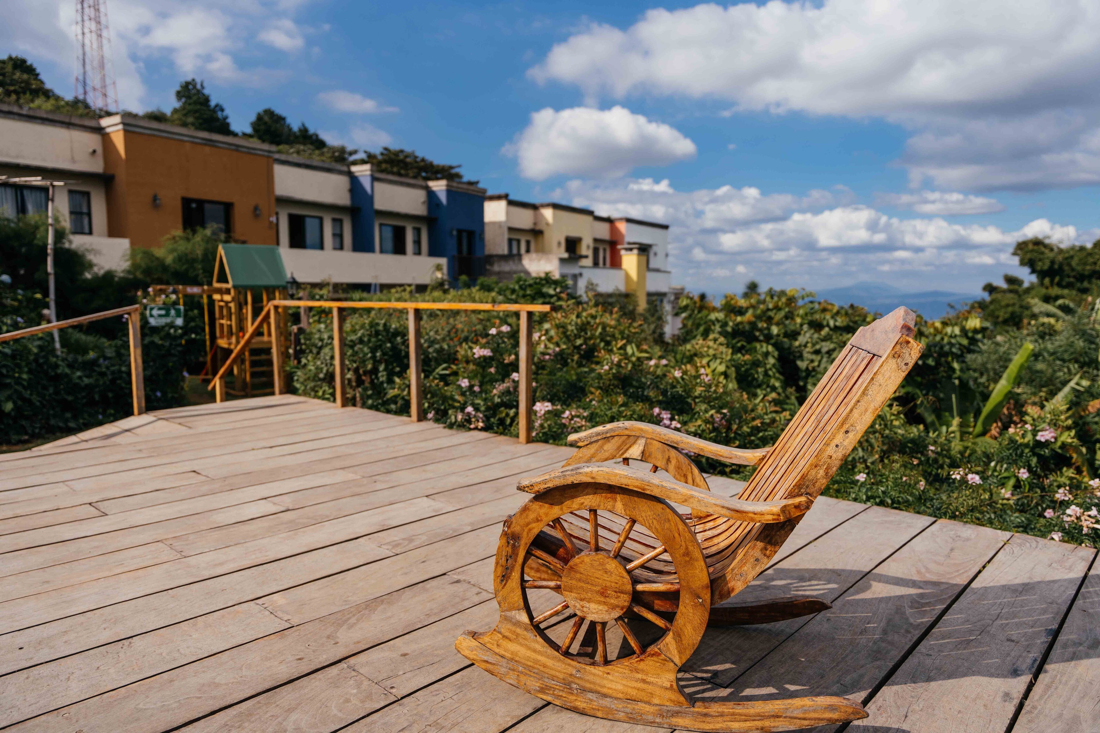 A wooden rocking chair on a wooden deck with a view of buildings and greenery in the background.