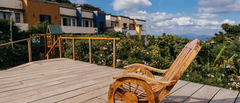 A wooden rocking chair on a wooden deck with a view of buildings and greenery in the background.