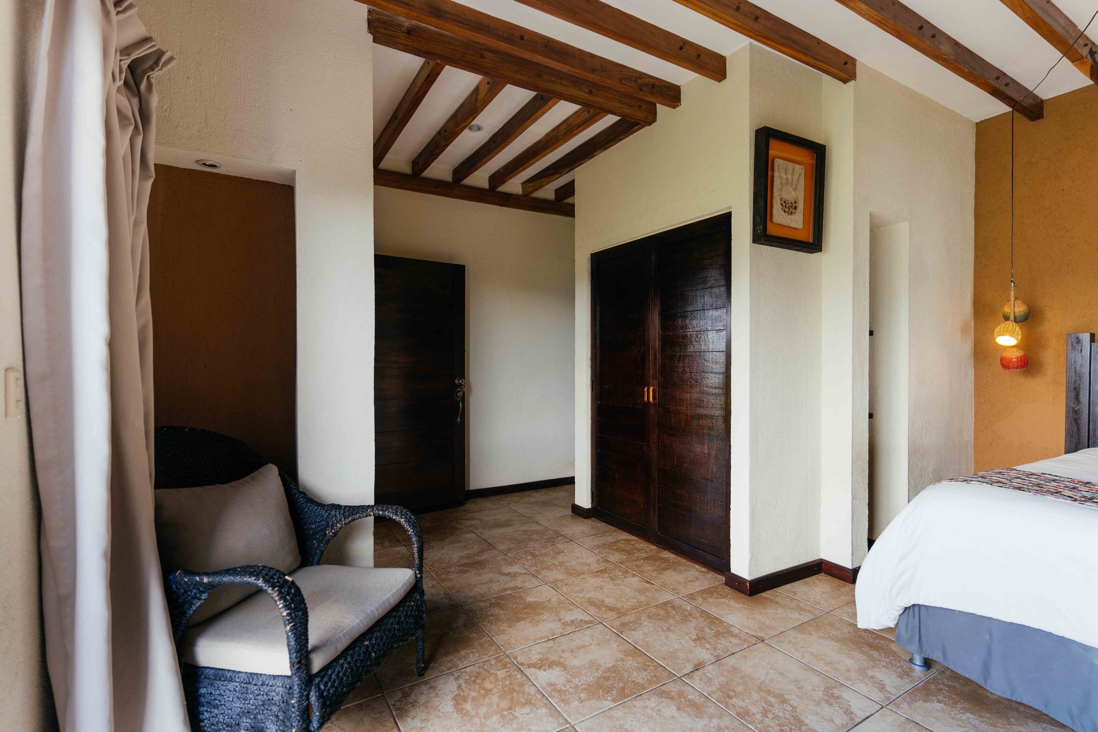 A hotel room with a bed, a chair, a door, and a ceiling with wooden beams.