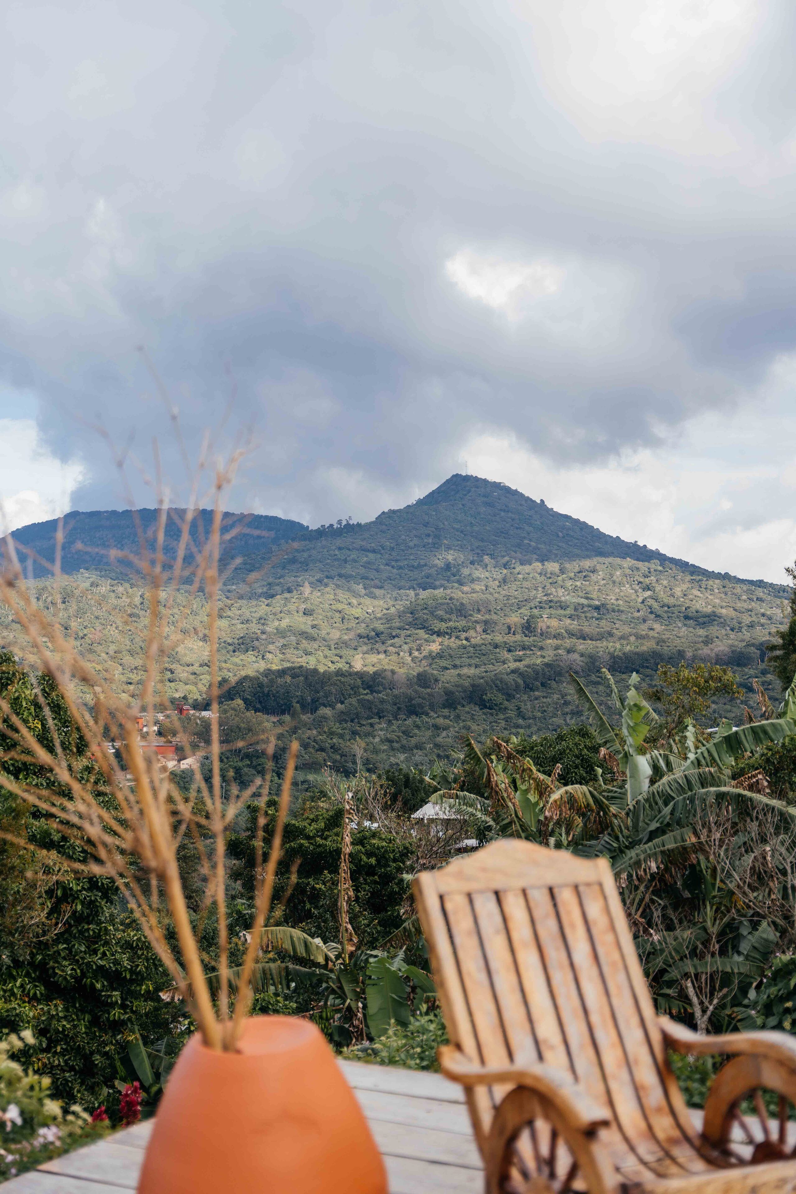 Une chaise en bois et un pot en terre cuite sont posés sur une terrasse en bois, d’où l’on peut admirer une montagne.