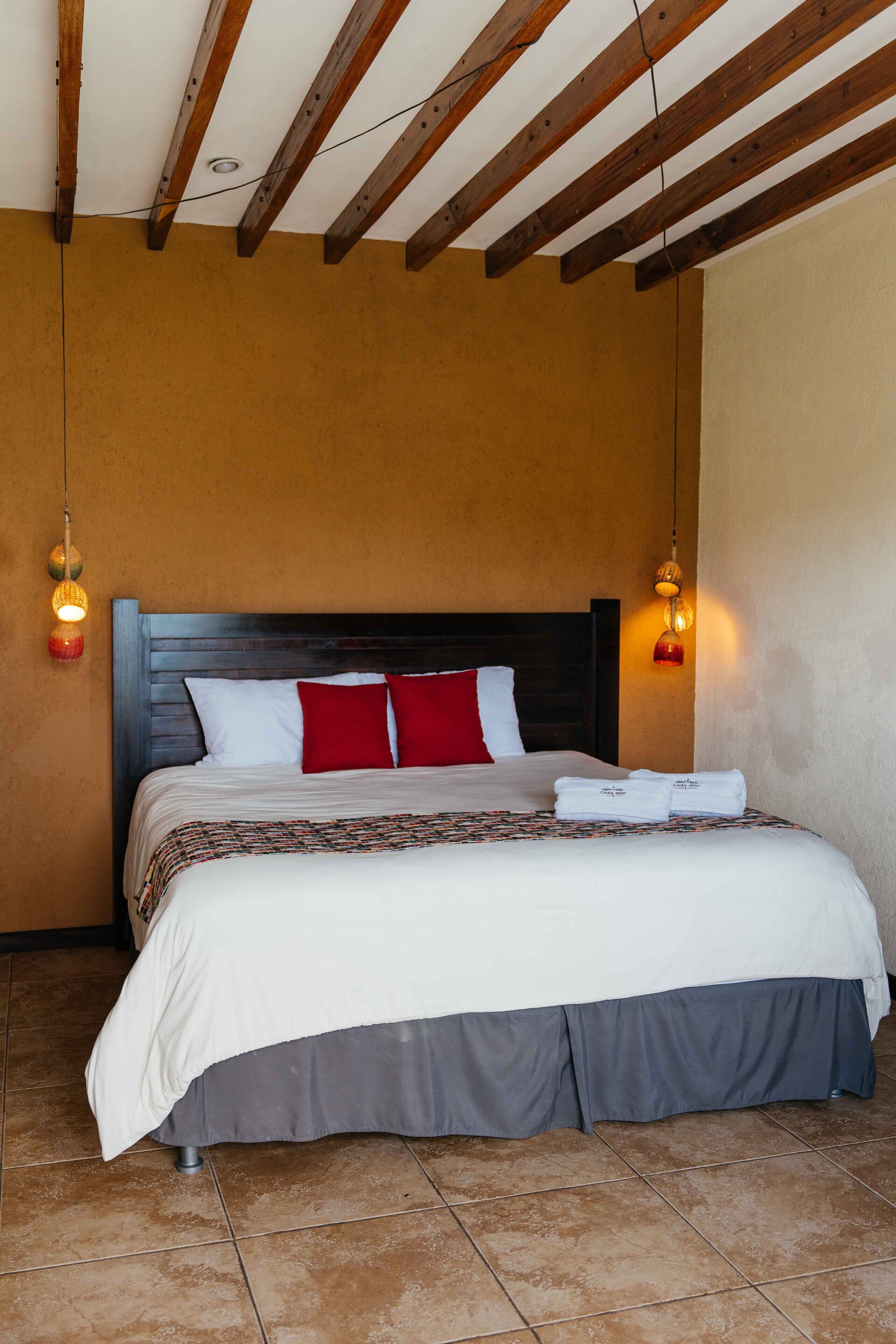 Une chambre d’hôtel avec un plafond en bois, un grand lit avec une literie blanche et rouge, et deux lampes fixées au mur.
