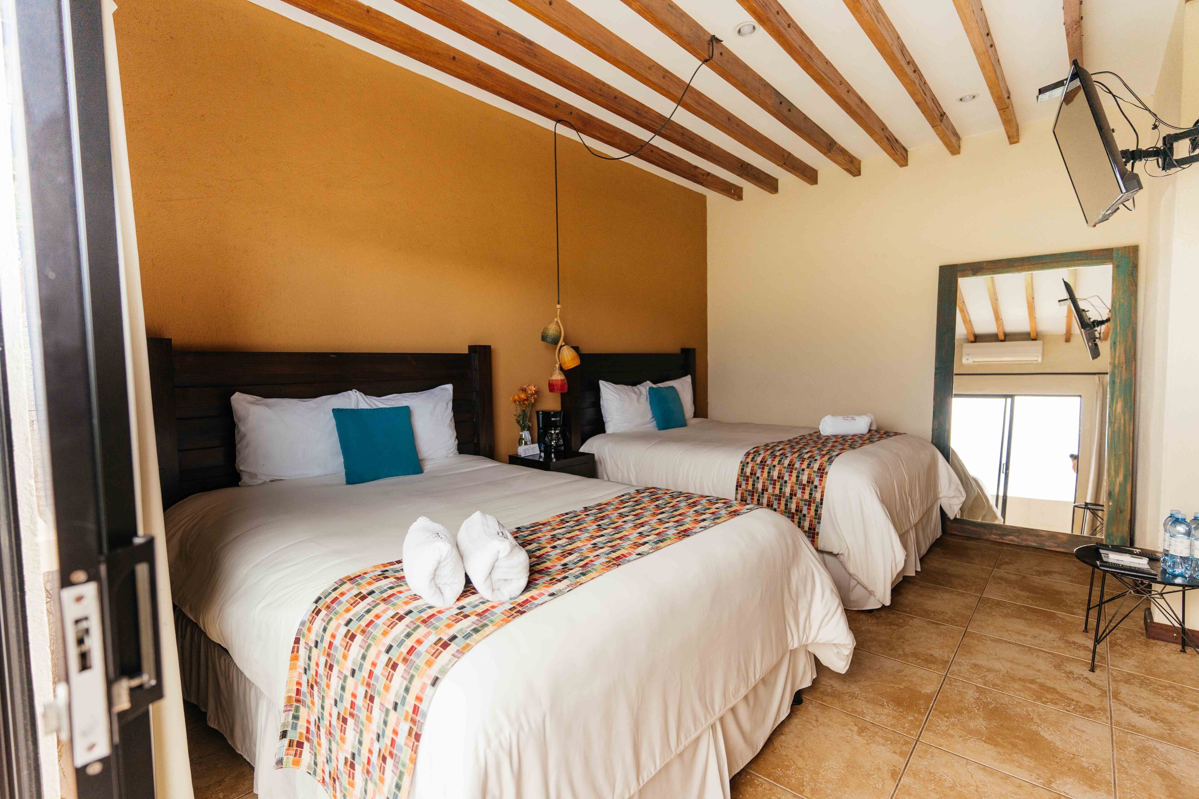 A hotel room with two beds, wooden ceiling beams, a small table with a lamp, and a window with an air conditioning unit.