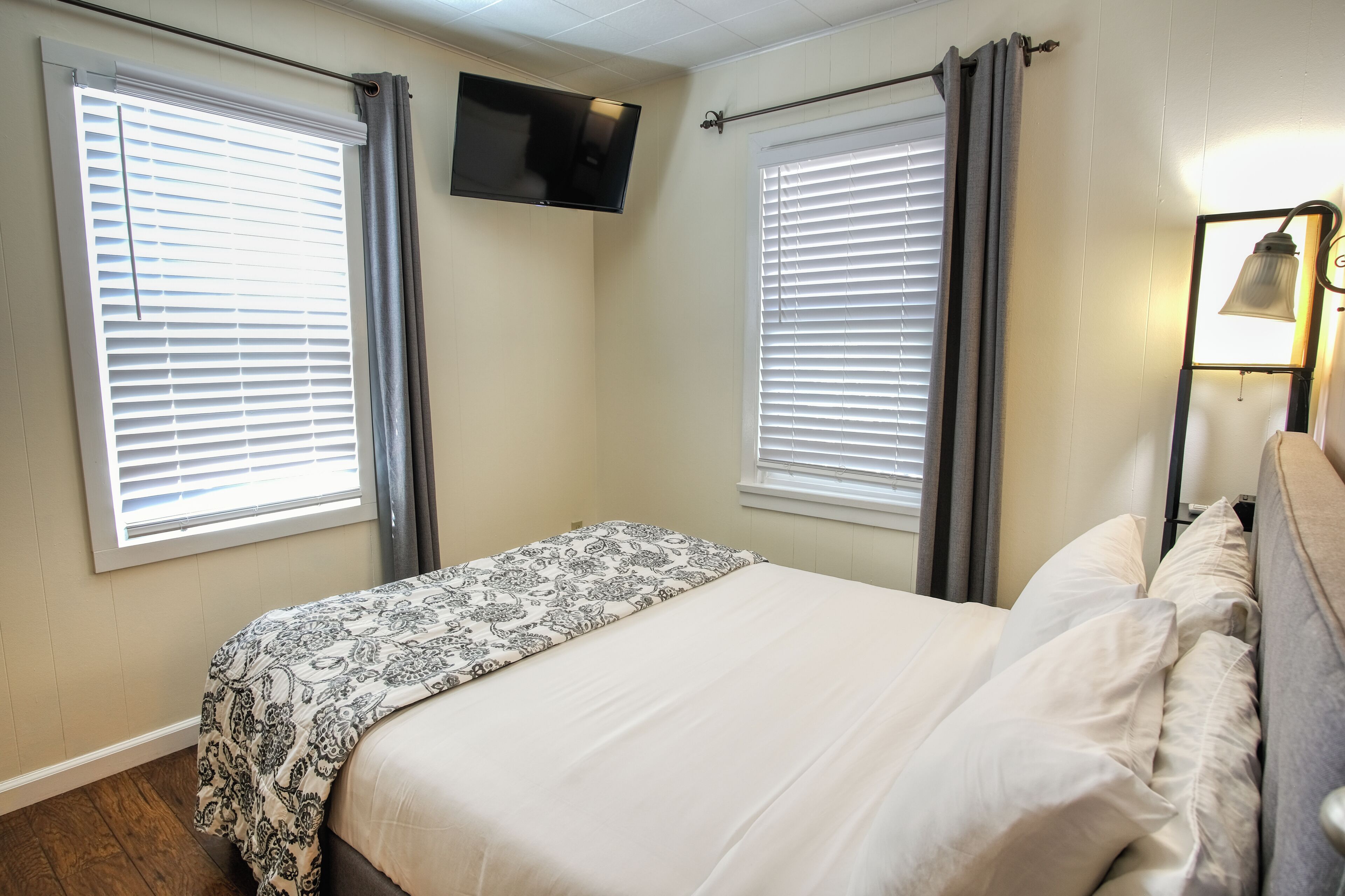 A bedroom with a bed, two windows with blinds, a flat-screen TV mounted on the wall, and a bedside table with a lamp.