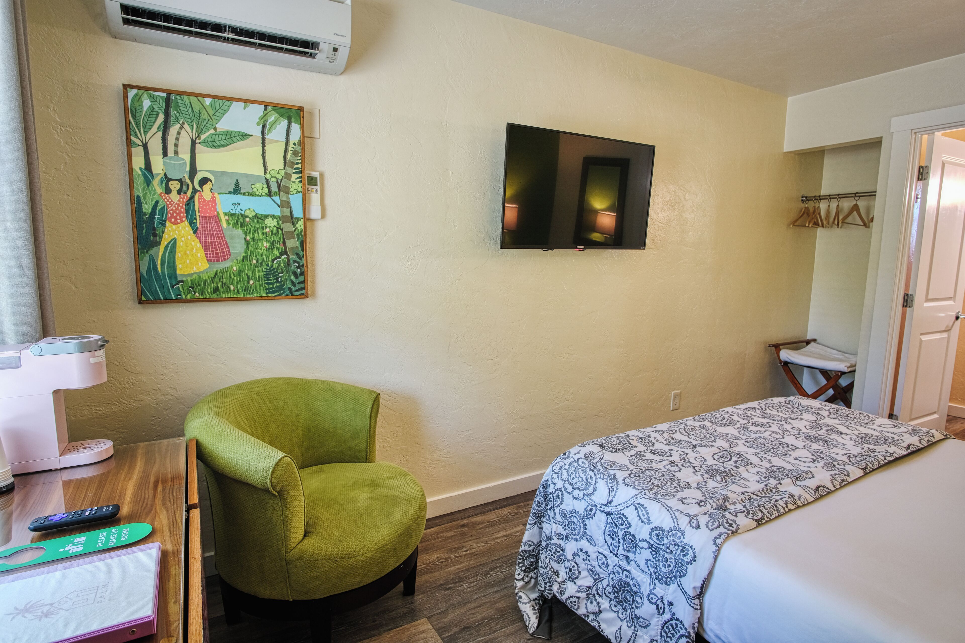A hotel room with a bed, a green armchair, a desk with a lamp, a painting on the wall, and a television.
