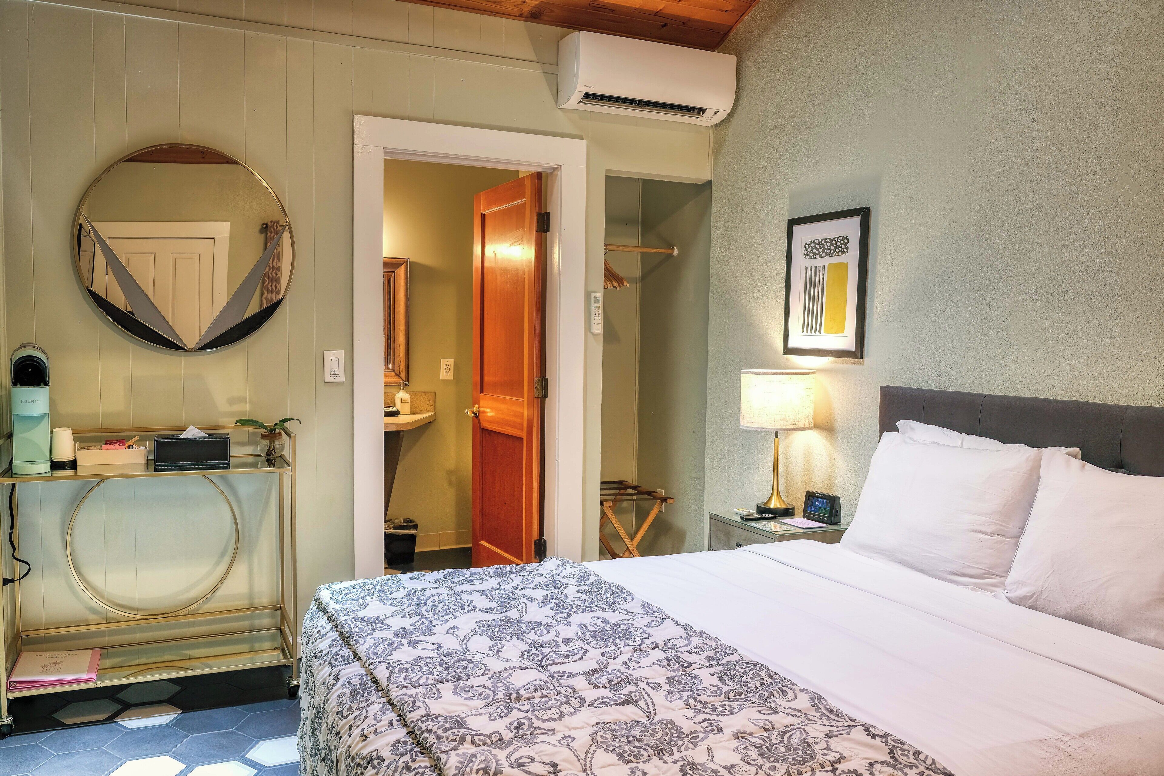 A hotel room with a bed, a round mirror, a bedside table with a lamp, a framed picture on the wall, and a door leading to a bathroom.