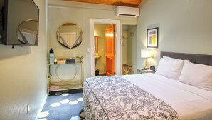 A hotel room with a bed, a mirror, a round mirror, a door leading to a bathroom, a nightstand with a lamp, and a framed picture on the wall. - Palm Cottages (Ashland)