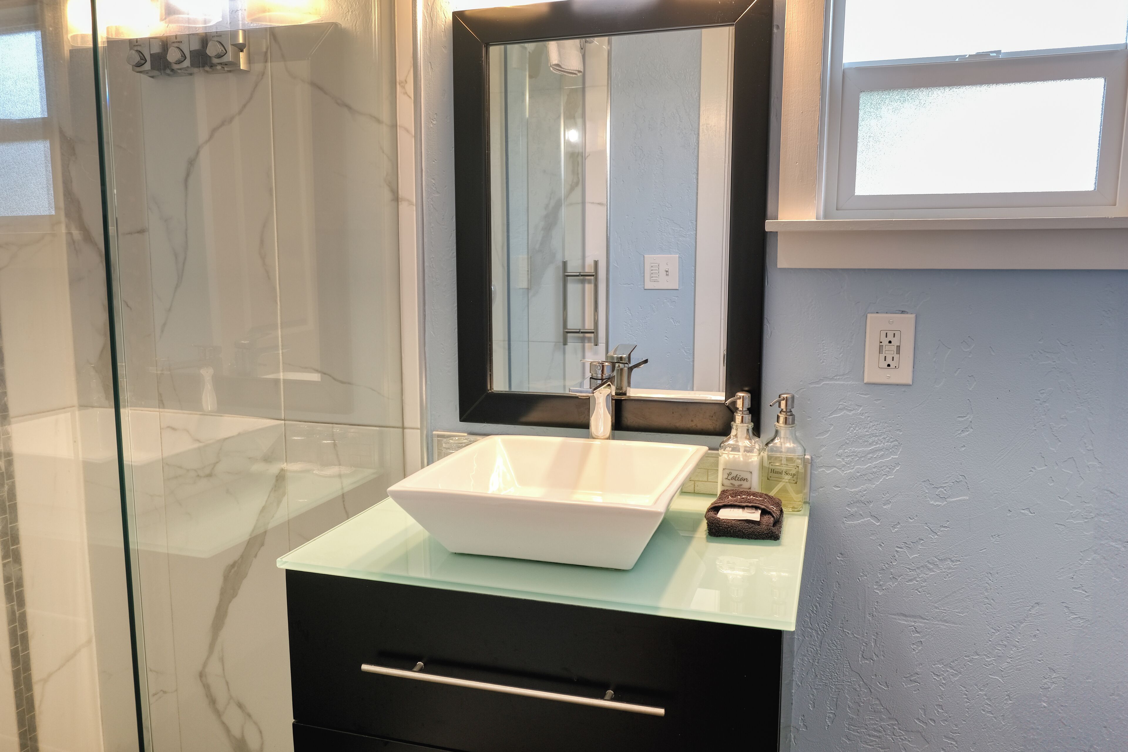 A bathroom with a glass-enclosed shower, a white sink with a marble countertop, and a mirror above the sink.