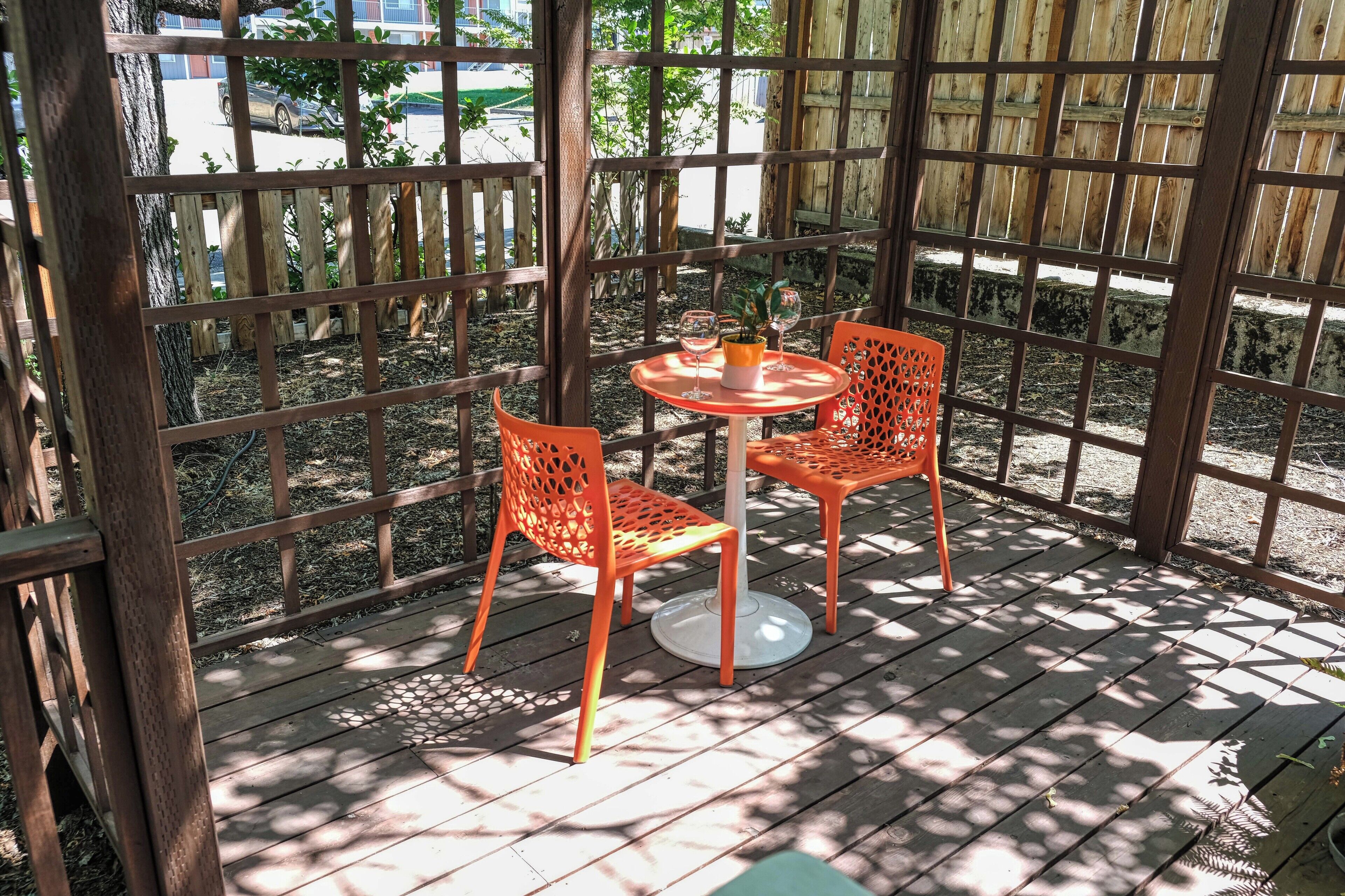 Une terrasse couverte avec une plateforme en bois, deux chaises orange et une petite table ronde sur laquelle se trouvent un verre et une bougie.