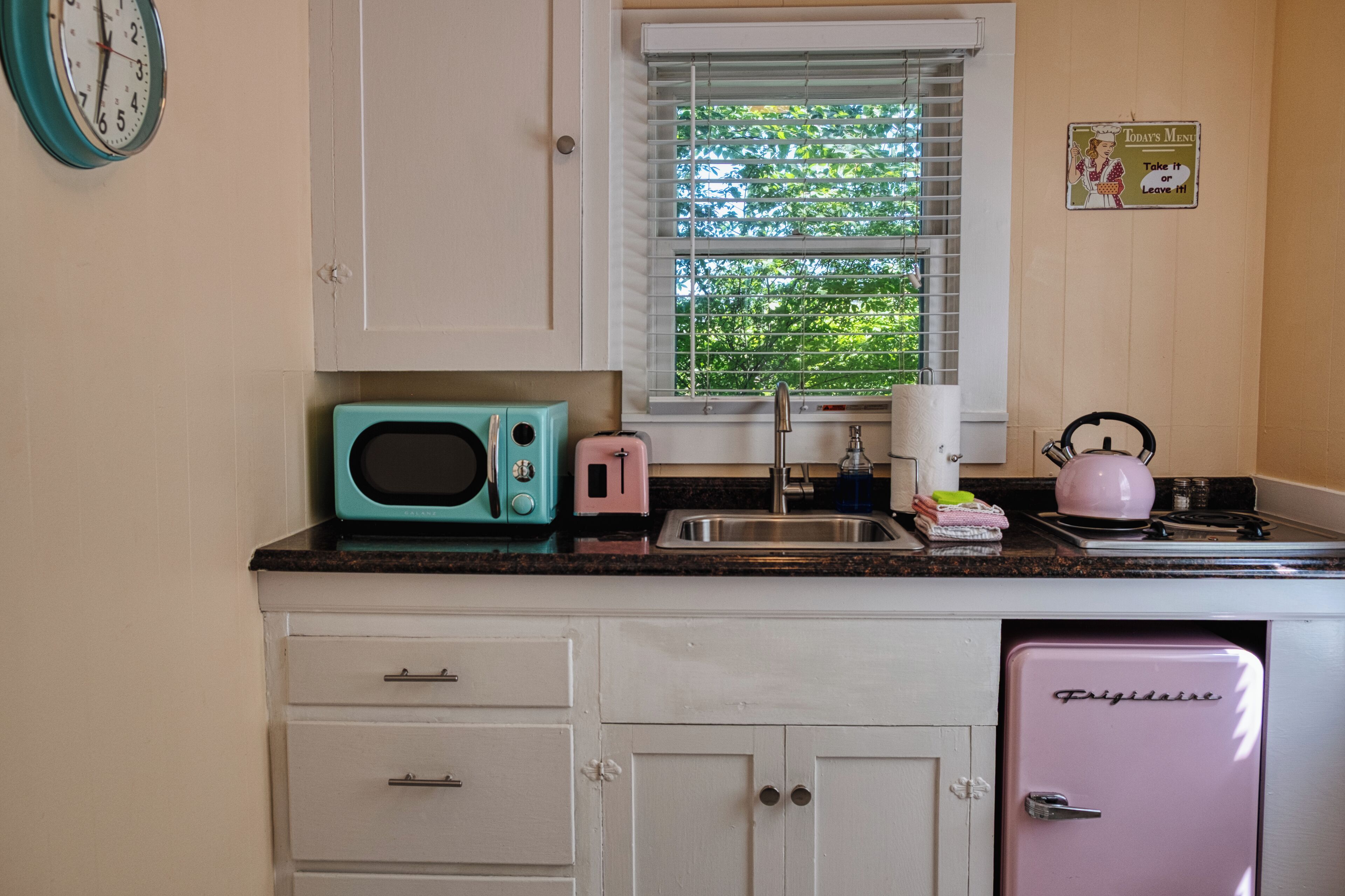 A kitchen with white cabinets, a turquoise microwave, a pink toaster, a sink, and a small refrigerator.