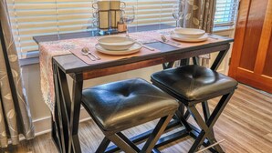 A dining table set with plates, cutlery, and glasses, with two leather stools and a window with blinds in the background. - Palm Cottages (Ashland)