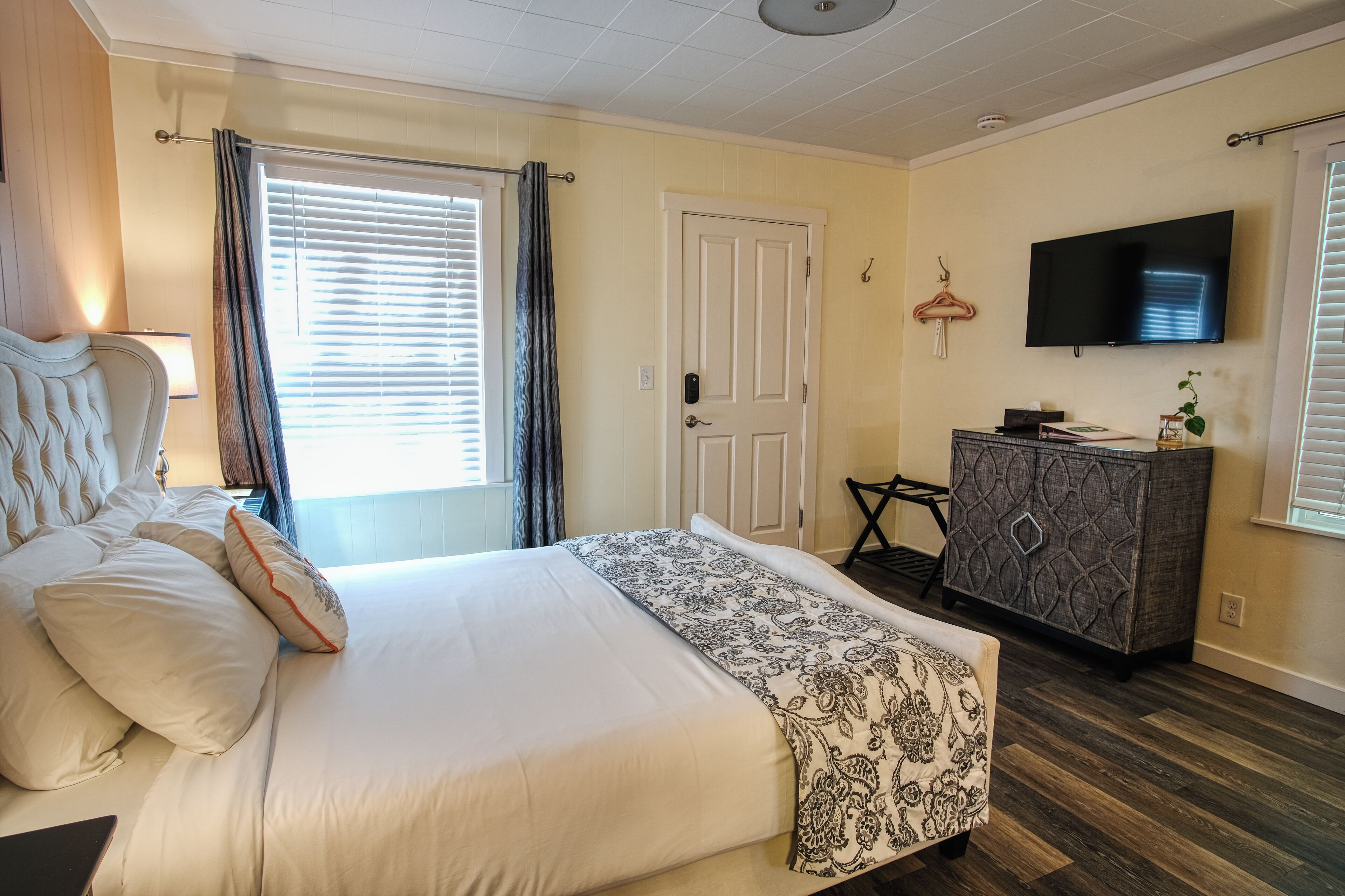 A hotel room with a large bed, a TV mounted on the wall, a dresser, and a window with blinds.