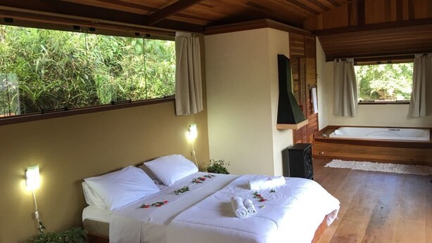 Ein Schlafzimmer mit Holzbalkendecke, einem Bett mit weißer Bettwäsche, einem Nachttisch, einem Fenster mit Vorhängen und Blick ins Grüne.