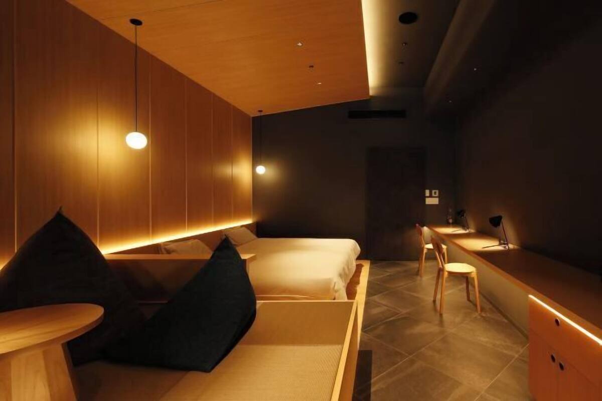 Quarto de hotel moderno com teto de madeira, duas camas com almofadas pretas, uma mesa redonda pequena e uma escrivaninha com abajur.
