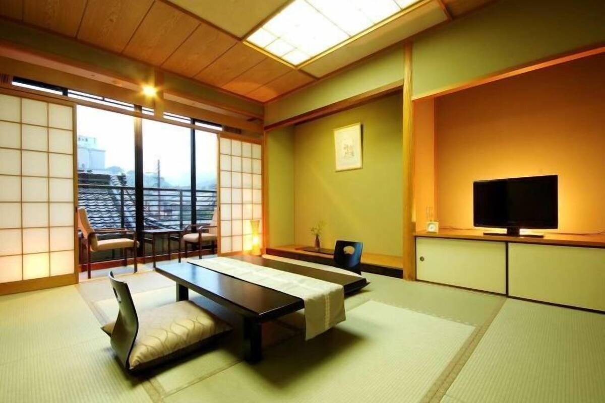 Um quarto japonês tradicional com tatames, uma mesa baixa, almofadas, uma televisão e vista panorâmica por grandes janelas.
