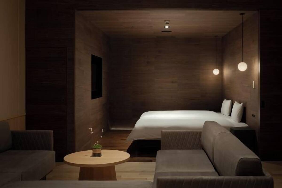 Quarto de hotel moderno com uma cama grande, um sofá, uma pequena mesa redonda e iluminação pendente.
