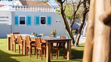 Un'area pranzo all'aperto con un tavolo e sedie in legno, un edificio bianco con persiane blu sullo sfondo e un albero in primo piano.
