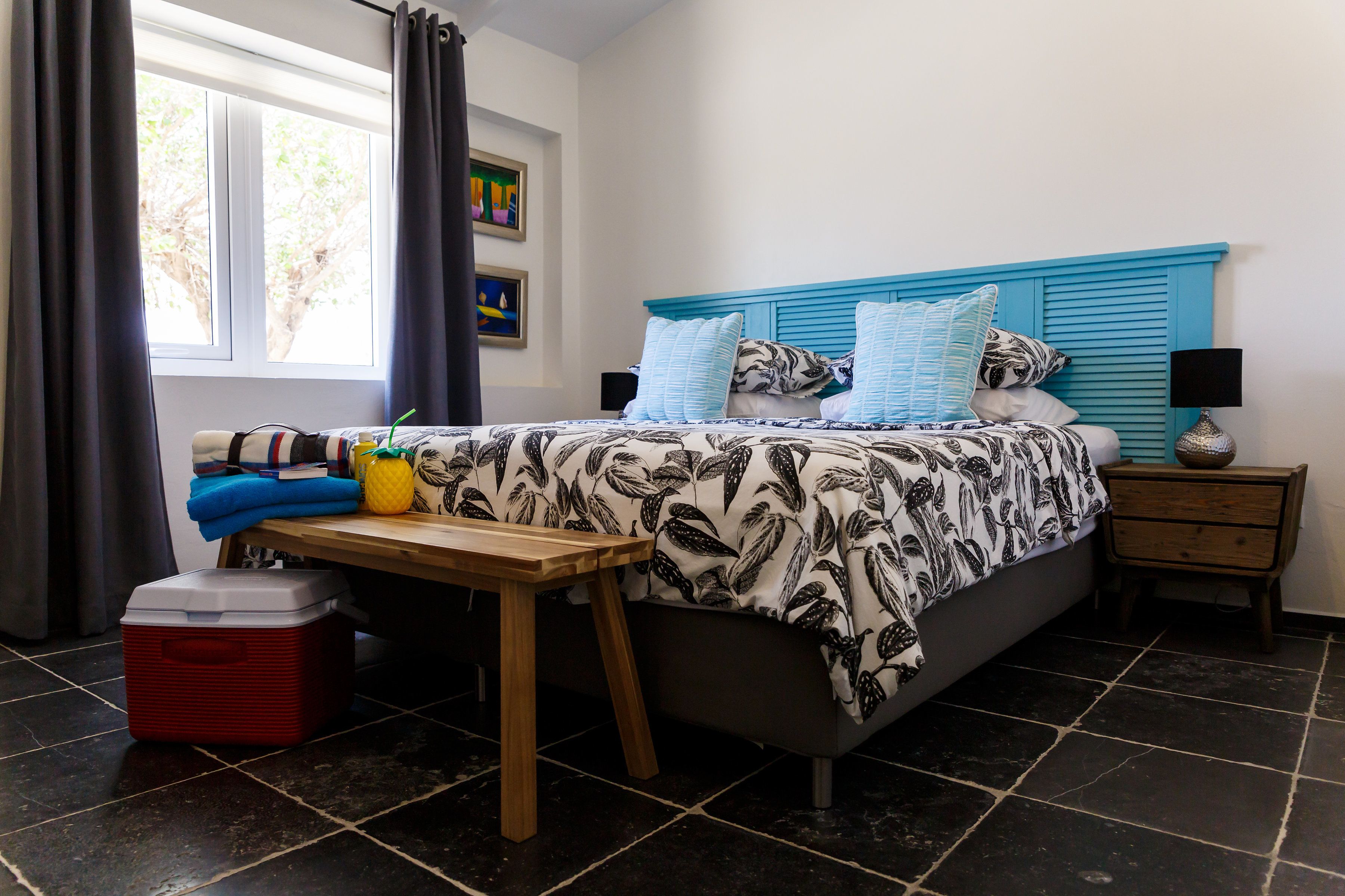 Un dormitorio con una cama, una mesita de noche, una mesa, un nevera portátil y una ventana con cortinas.