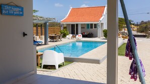 A pool area with a tiled deck, a covered seating area, and a house with a red-tiled roof in the background. - Aruba Boutique Apartments Adults Only (Noord)