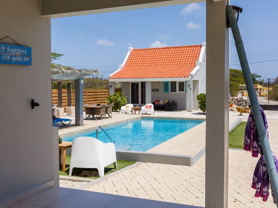 A pool area with a tiled deck, a covered seating area, and a house with a red-tiled roof in the background.