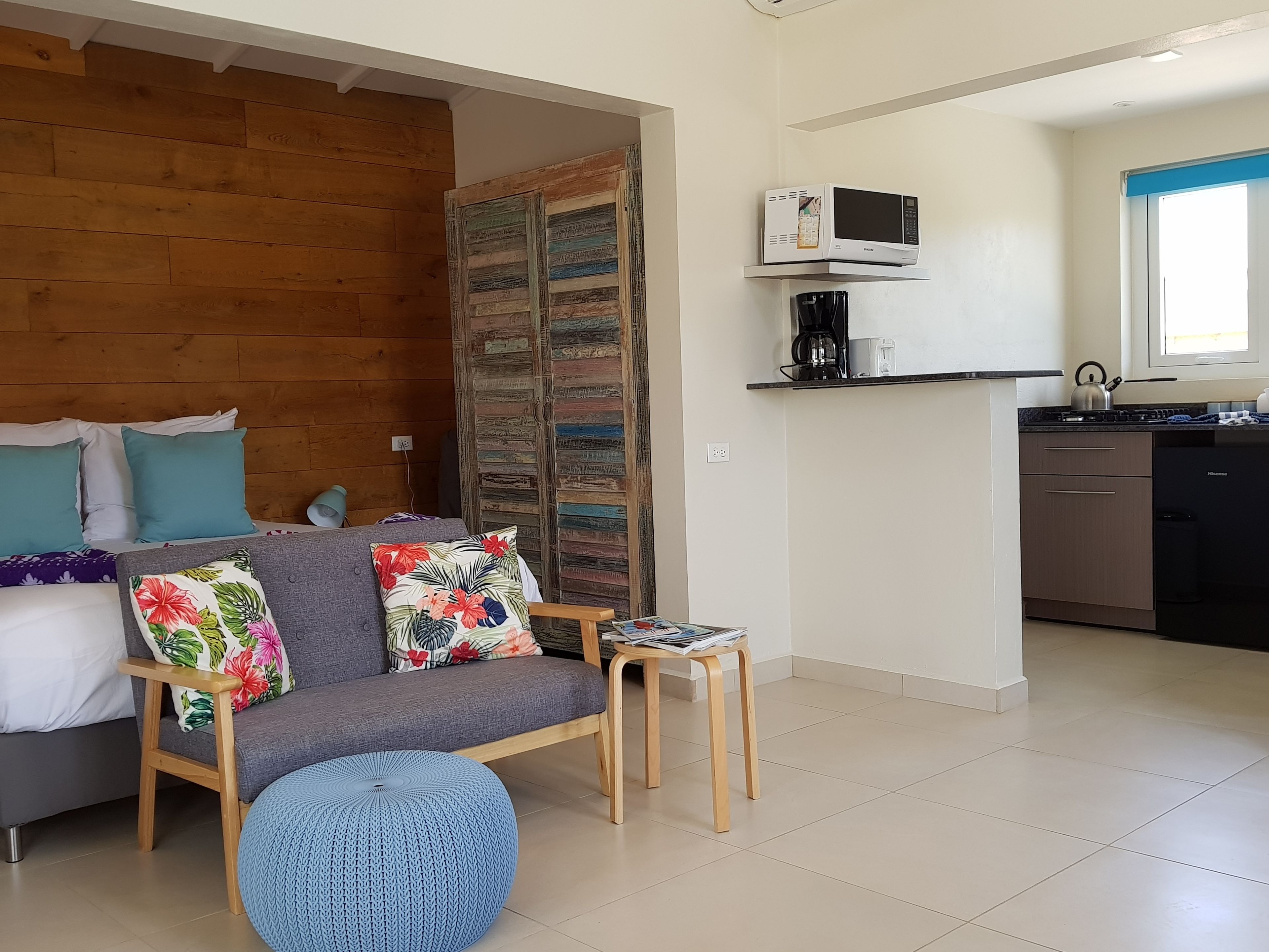 A cozy living room with a sofa, a small table, and a blue ottoman. A wooden partition separates the living room from a kitchen area with a microwave and a kettle.