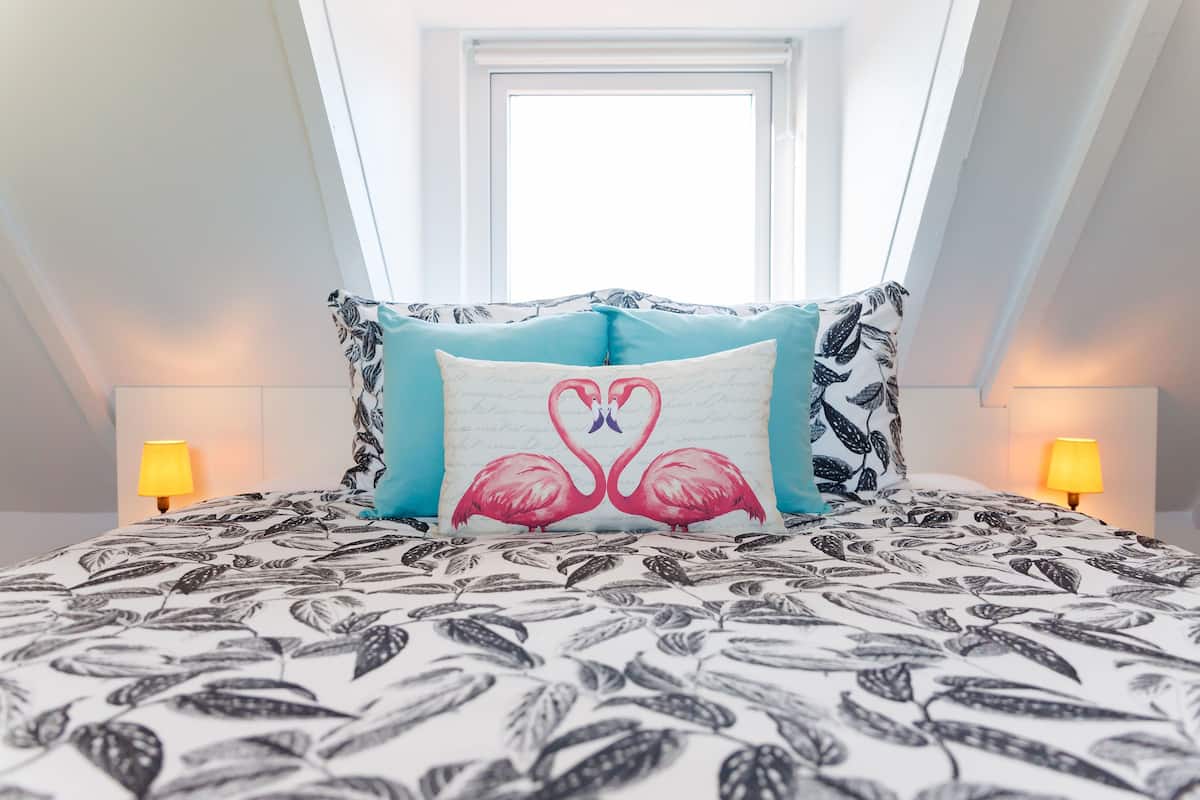 A bedroom with a bed featuring a leaf-patterned bedspread, a pillow with flamingos, and a skylight above.