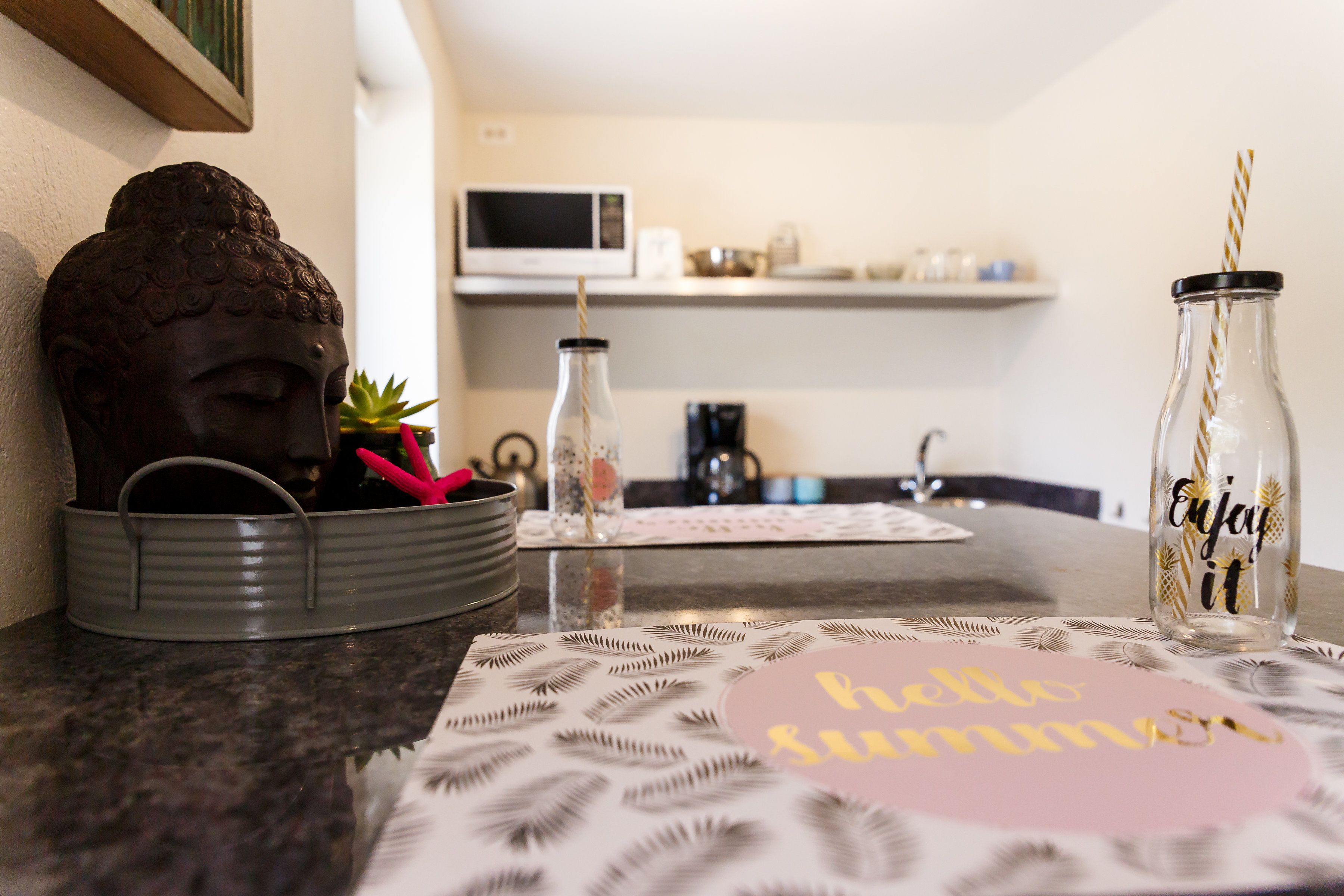 Una encimera de cocina con una estatua de Buda, una botella de vidrio con una pajita y un mantel individual con la frase 'hello summer'.
