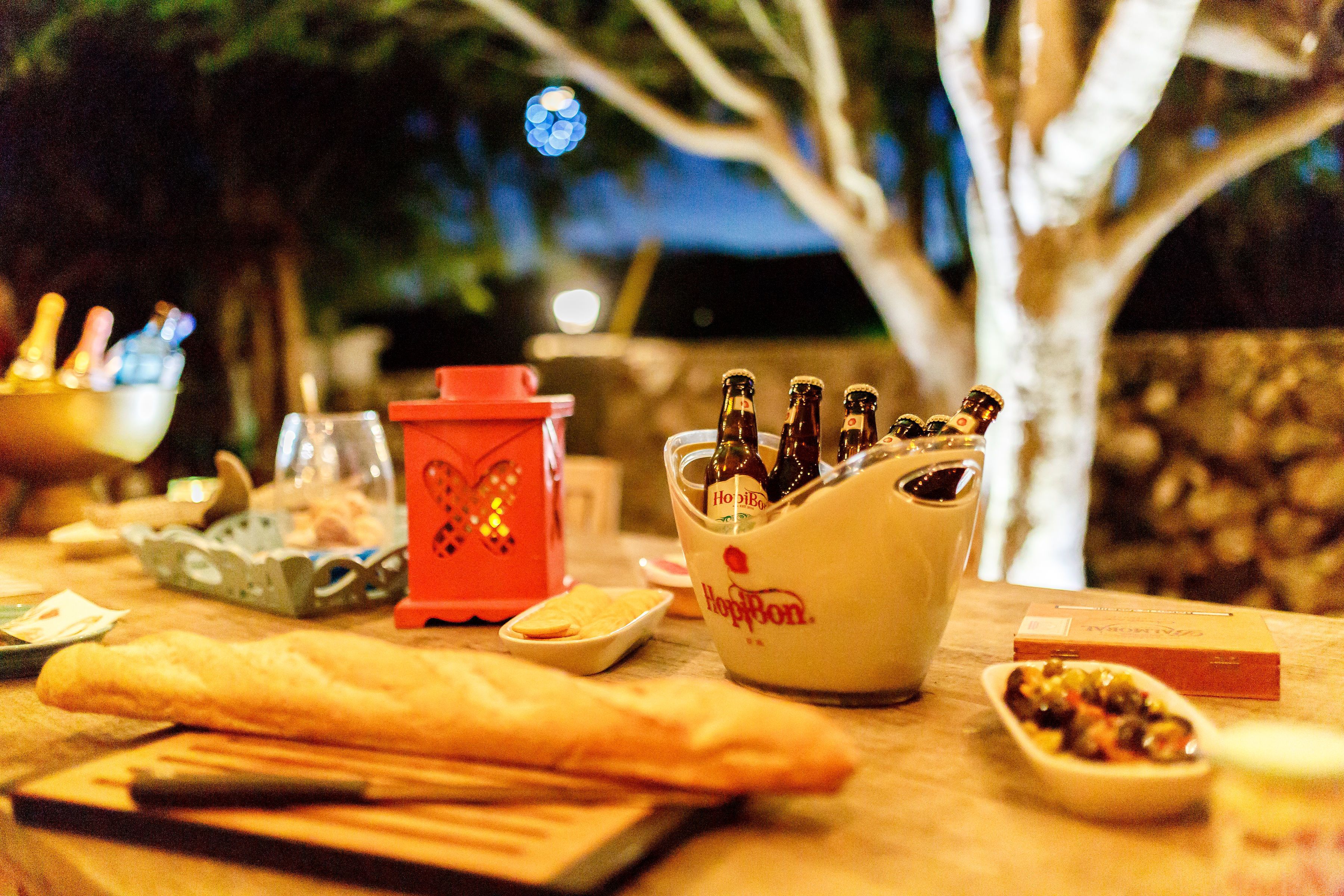A table set with food and drinks, including a bowl of olives, a red container, and bottles of beer.