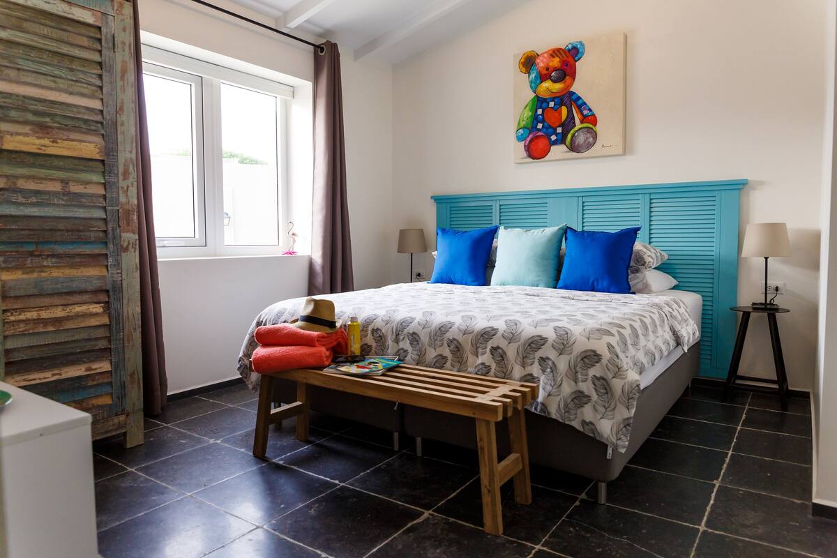 A bedroom with a bed, a bench, a window, a painting, and a lamp.