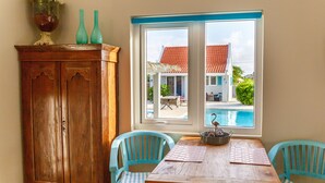 A dining area with a wooden table and two turquoise chairs, a wooden cabinet, and a window with a view of a house and a swimming pool. - Aruba Boutique Apartments Adults Only (Noord)
